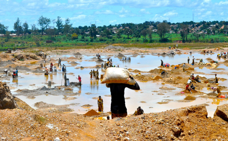 Zahlreiche Menschen arbeiten unter freiem Himmel im Kupferabbau in einer schlammigen, wasserbedeckten Landschaft im Kongo. Eine Person trägt einen schweren Sack auf dem Rücken, während andere in kleinen Gruppen graben oder schürfen. Im Hintergrund ist eine grüne Vegetation und eine Siedlung zu sehen.